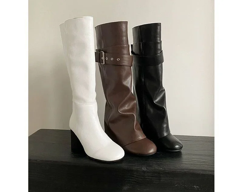 Buckled Knee-High Riding Boot — A knee-high leather riding boot with a buckled belt strap across the top of the shaft and a fold at the ankle, shown in brown, milky white and black.