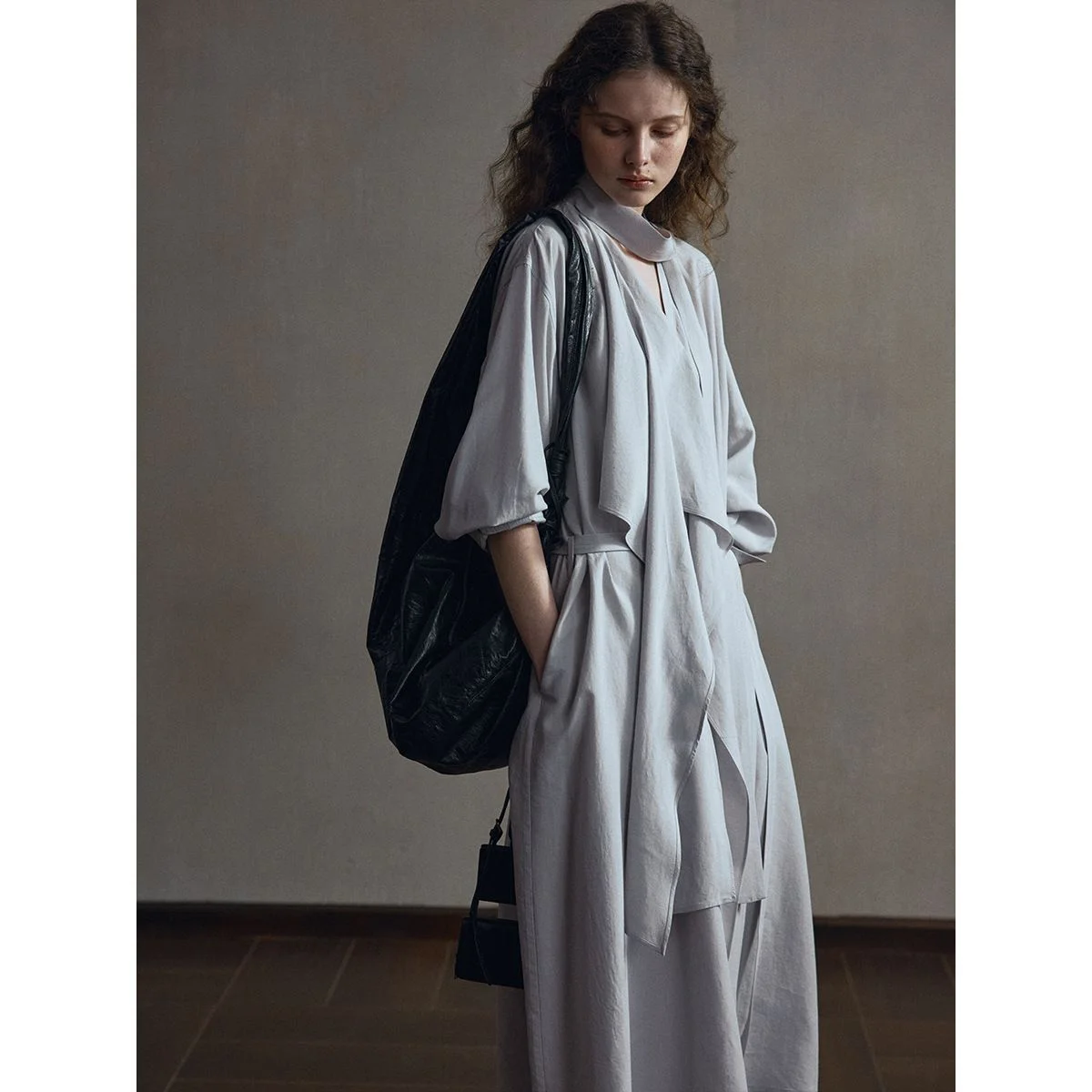 Lyocell-Linen Trench Dress — A long loose light grey lyocell-linen shirt-coat with a tie belt, worn by a model against a grey studio wall